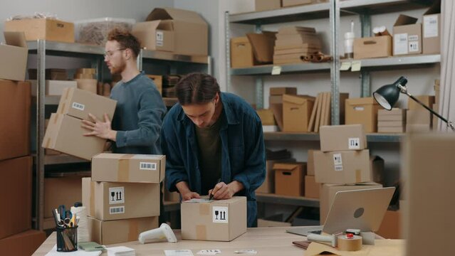 Caucasian postman and his male colleague putting stickers at the cupboard box while preparing delivery parcel box. Post service and small business concept.
