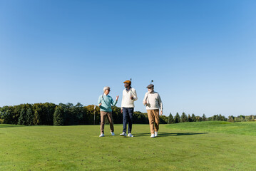 senior interracial friends walking with golf clubs on field.