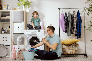 Father and son are putting clothes in wash. A small child sits on the washing machine helping dad,...