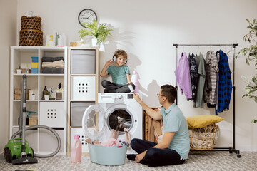 Father and son are putting clothes in wash. A small child sits on the washing machine helping dad,...