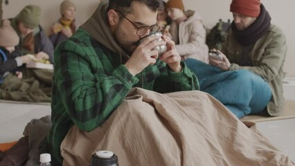 Slowmo portrait of bearded refugee man in eyeglasses looking at camera after drinking hot tea from thermos cup sitting on floor in cold asylum with other refugees