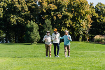 senior multiethnic friends smiling and walking with golf clubs on green field.