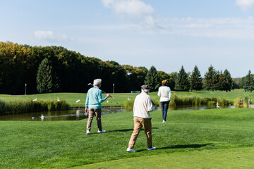 back view of senior multiethnic men with golf clubs walking near pond with swans.