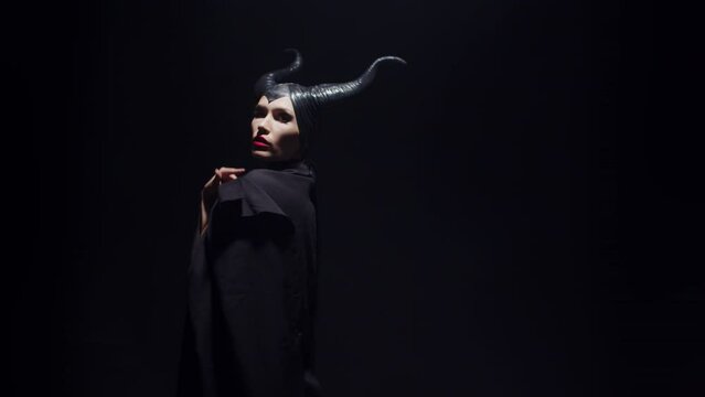 Woman in witch costume waves cloak standing at studio on black background, front view. Woman in sorceress costume opens cloak