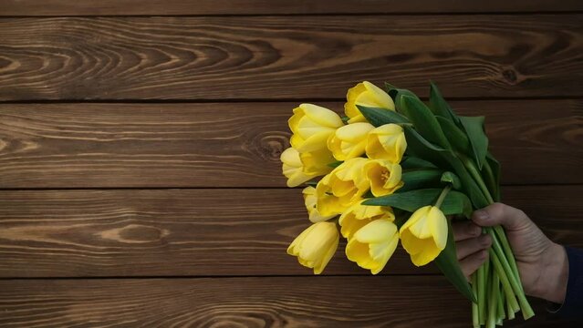 a man gives a beautiful bouquet of fresh flowers. a man's hand holds out yellow tulips on a wooden background. the concept of a gift and congratulations for Mother's Day, Valentine's Day, Women's Day.