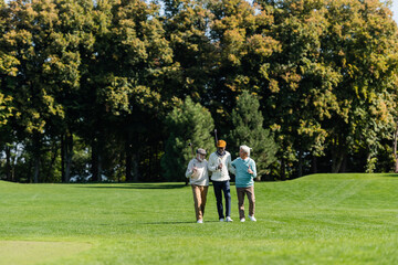 senior multiethnic golfers walking with golf clubs on green field near trees.