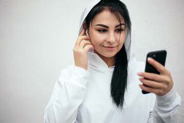 Woman dressed white hoodie hold phone listening music with wireless headphones while training running jogging doing sport at the street in the morning. Sporty female athlete switching the song
