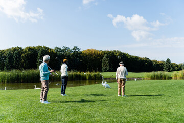 back view of senior multiethnic friends with golf clubs standing on green lawn near pond.