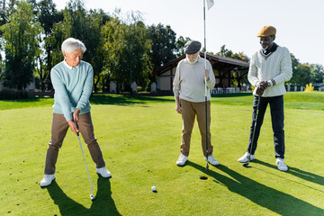 Obraz premium senior man in flat cap holding flag stick near hole and multiethnic friends.