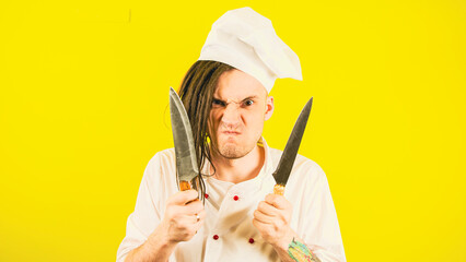 Young insane man dressed as chef with knives on yellow background. Crazy male cook in white hat and shirt posing with kitchen knives.
