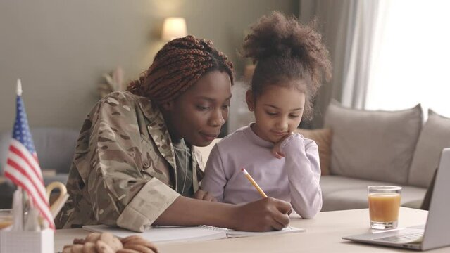 Waist up slowmo of young Black woman with dreadlocks in military uniform and her preschool daughter studying together at desk in living room, solving math examples