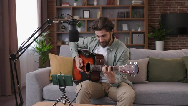 Young man recording guitar lessons for students. Online music class with distant teacher on mobile device. Tutor teaching music through smartphone. Online acoustic guitar courses, distance education.