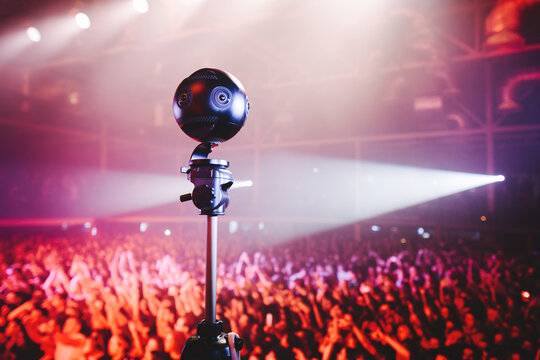 Professional 360 camera at music concert on a tripod recording performance on video.
silhouettes of crowd in front of bright stage lights.