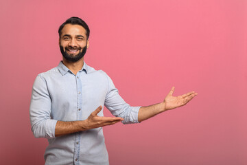 Handsome Indian guy points palms at empty copy space isolated on pink background. Cheerful...