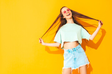 Portrait of young beautiful smiling female in trendy summer jeans skirt. carefree woman posing near yellow wall in studio. Positive model having fun indoors in sunglasses. Cheerful and happy