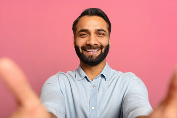 Portrait of bearded Indian handsome man taking selfie, looking at camera POV, multiracial brown-haired guy recording himself. Indoor studio shot isolated on pink background