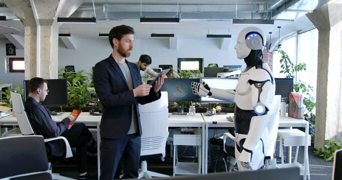 Man programming robot with tablet computer in hands in office. Male developer checking work of robotic by device. Coworking space. Humanoid moving and executing instructions and orders.