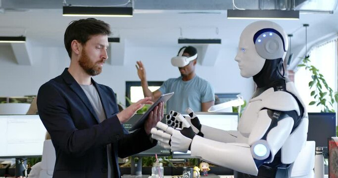 Man programming robot with tablet computer in hands in office. Male developer checking work of android by device. Humanoid moving and executing instructions and orders. Artificial intelligence startup