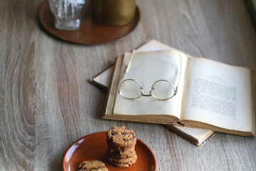 Stack of chocolate chip cookies, open book and reading glasses, lit candle and vase with flowers on the table. Hygge at home. Selective focus.