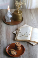 Stack of chocolate chip cookies, open book and reading glasses, lit candle and vase with flowers on the table. Hygge at home. Selective focus.