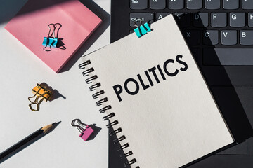 Word POLITICS on a notepad lying on keyboard of a laptop next to pink sticky notes, black pencil and paper clips.