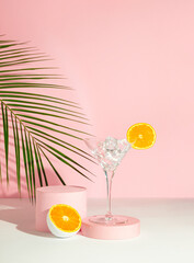 Martini glass with ice and orange fruit, tropical palm leaf and podium on pink background. Summer...