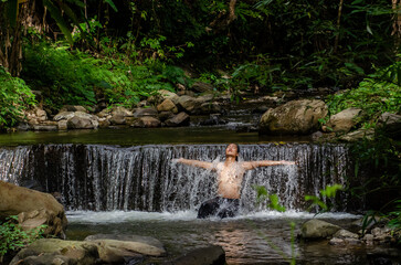 Asian man relaxing with freedom open arms in waterfall in tropical nature. Wellness spa concept in holiday nature.