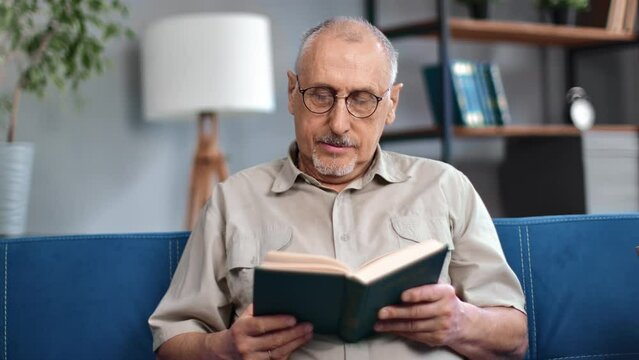 Happy mature old 70s man gray haired man reading interesting paper book resting on couch retirement 
