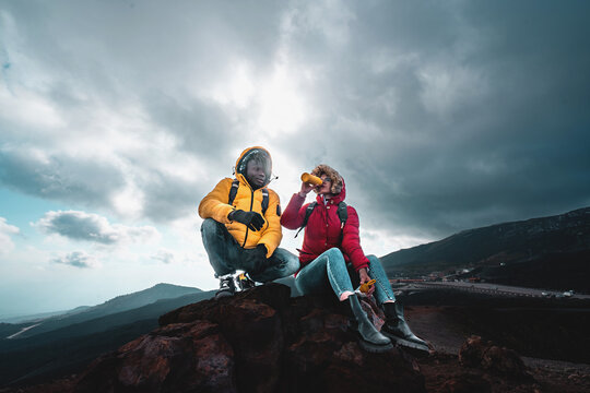 Biracial couple of young people relaxing on the top of the mountain sitting on the lava stones and drinking water in a plastic-free bottle - tourism, wanderlust and lifestyle concept