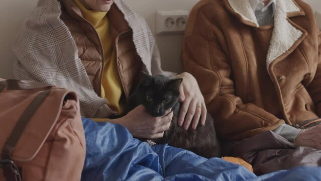 Cropped slowmo of mature refugee woman wearing old warm clothes sitting with other refugees indoors in empty room petting homeless black cat
