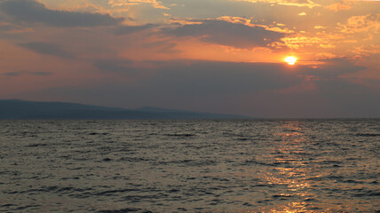 Fototapeta premium Sunset over the sea in pink bronze tones, Brela, Croatia. The orange sun sets behind the clouds, the sun rays are reflected on the surface of the sea on a summer evening