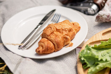 Croissant on a white plate with a fork and knife. Romantic outdoor picnic.