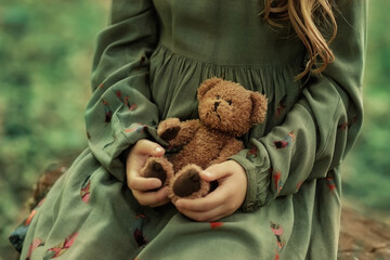 A girl in a green dress holds a teddy bear in her hands. Green leafy background.