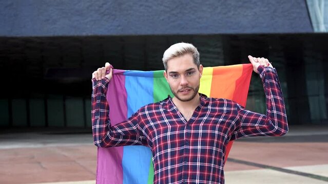 Young gay man holding rainbow flag outdoor at pride parade while smiling on camera
