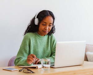 Young African-American female working on laptop in headphones while taking notes in notebook