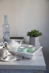 A blue transparent water bottle, teapot, the two green cups, tray on a white wooden tray on the kitchen table.