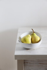 Two green pears in a white faience bowl on the edge of a beige kitchen table. Scandinavian style. Minimalism. Place for text.