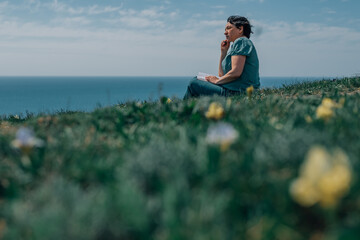 Naklejka na ściany i meble adult woman thoughtfully writes plans diary on a sunny day on a mountain against backdrop of the sea. pensioner sits on the grass barefoot in summer, spring, with a notebook and a pen