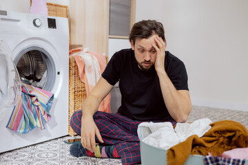 Guy sits on the laundry room floor with the washing machine open, throwing all the clothes into the...