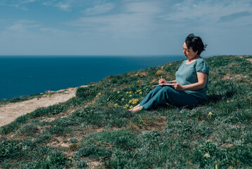 Naklejka na ściany i meble adult woman thoughtfully writes plans diary on a sunny day on a mountain against backdrop of the sea. pensioner sits on the grass barefoot in summer, spring, with a notebook and a pen