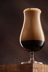 Glass of porter beer with overflowing foam head on the corner of a wooden table. Shallow depth of field.