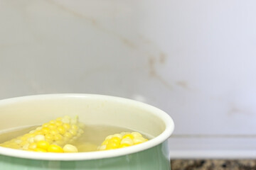 Healthy corn on the cob (sweet corn) boiling in green pot of hot water in bottom left of frame, white marble wall in background