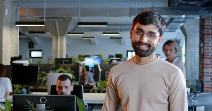 Portrait shot of Arabian successful handsome man looking at camera and smiling in office. Indoor. Cheerful male in glasses standing in comfortable modern coworking space.