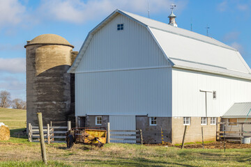 A small white dairy barn with a concrete silo and a rusty manure spreader in the foreground. © Margaret Burlingham