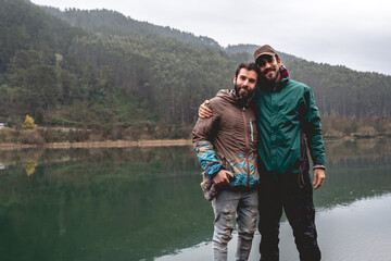 Two happy, handsome, young and bearded brother with cap and jackets smiling in a wood dock with view of lake in the mountains with green forest, mist, reflections in the water and cloudy sky