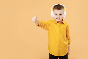 Portrait of funny clever school boy with headphones in yellow shirt. Yellow studio background. Education. Looking, smiling and shows a thumb up at camera