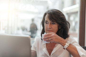 Smiling woman sitting in cafeteria holding coffee mug and working on laptop. Businesswoman checking email on laptop. Beautiful middle aged woman and using laptop at cafe while drinking a cup of tea.