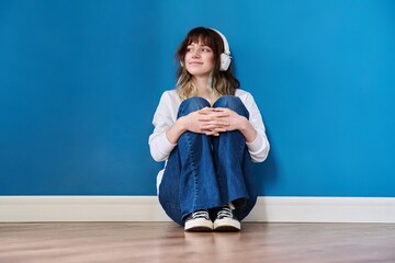 Portrait of teenage girl in headphones sitting on floor on blue background