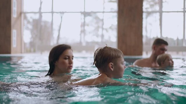 happy family is swimming together in large pool of wellness or hotel, parents and children are bathing in warm water