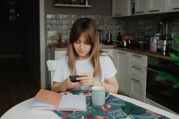 A young pretty woman is sitting at a table in the kitchen with a cup of coffee and a diary, using phone